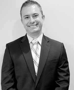 The image is a photograph of a man standing against a plain wall. He is wearing a dark suit and tie, with a light shirt and a striped tie. His expression is neutral and he appears to be posing for the picture.